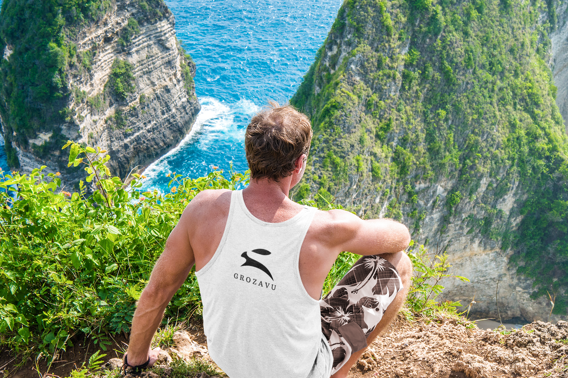 Back view of man in white Grozavu premium cotton tank top with rabbit logo, seated on a coastal cliff above the blue ocean