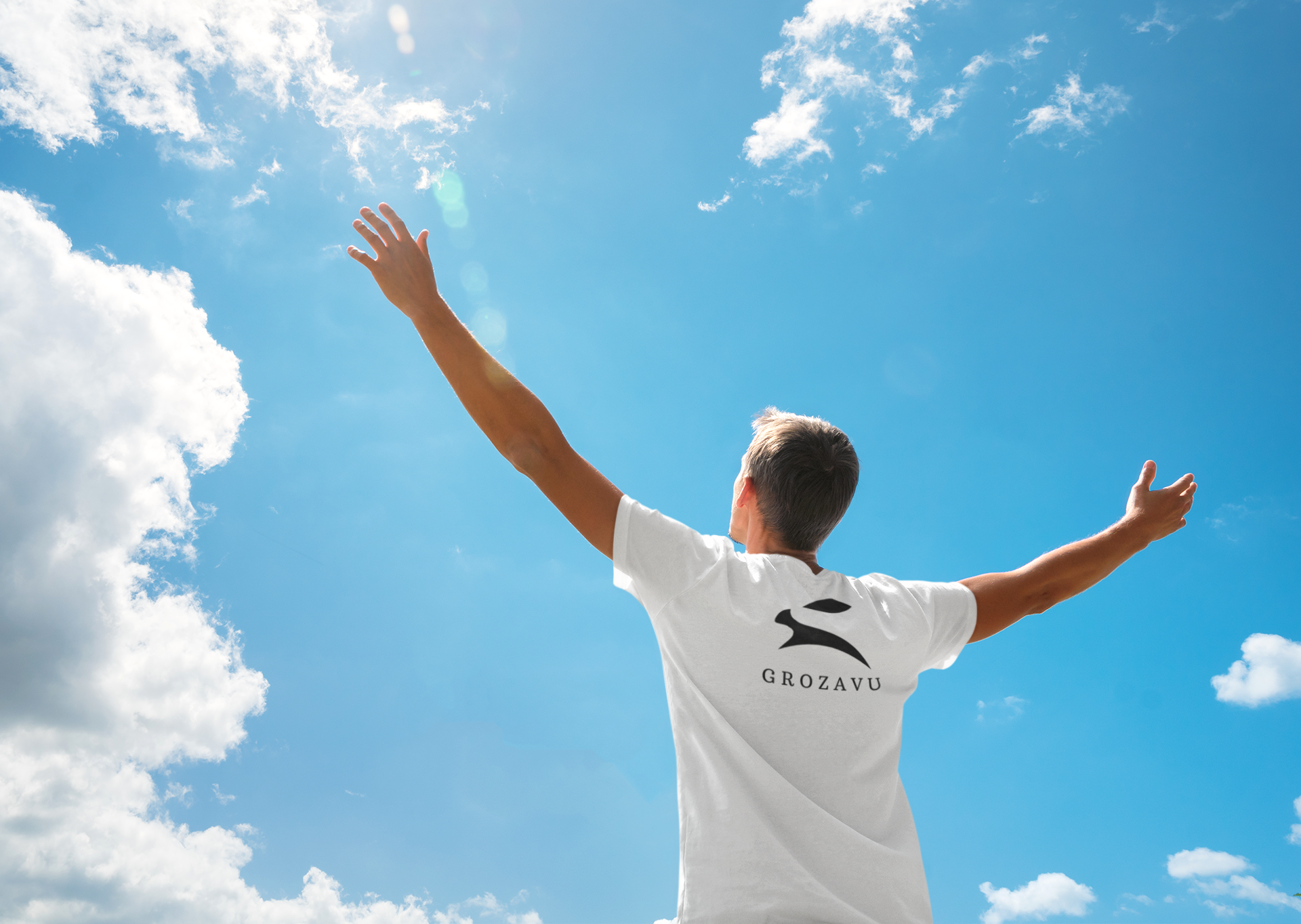 Back view of man in white Grozavu premium cotton T-shirt with rabbit logo, arms raised to blue sky, outdoor lifestyle