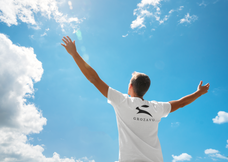 Back view of man in white Grozavu premium cotton T-shirt with rabbit logo, arms raised to blue sky, outdoor lifestyle