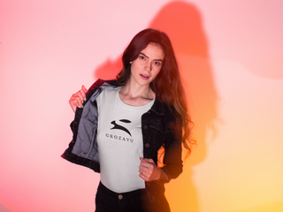 Woman revealing white Grozavu T-shirt with black rabbit logo under a denim jacket, pink–orange gradient studio background