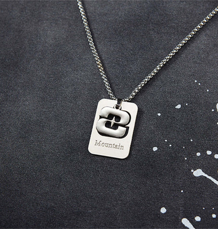 Close-up of a silver pendant necklace with a rectangular tag engraved with the word "Mountain" against a paint splatter background.